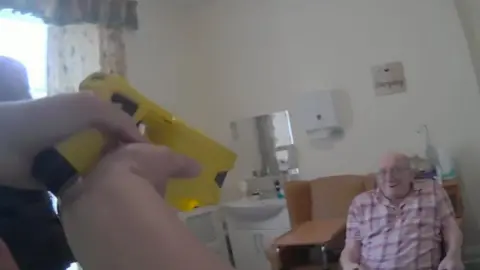 Independent Office for Police Conduct Donald Burgess sits in a chair beside his bed in a care home. He is wearing a red and white checked shirt. One police officer is visible to the left side of the picture and the hands of another, pointing a yellow taser at him, are also visible to the left side of the screen.