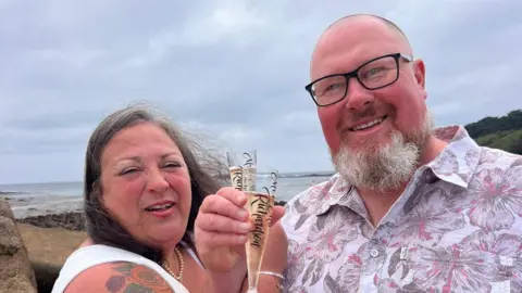 Julia Collins is pictured wearing a white wedding dress as she holds a glass of Champagne next to her new husband, Teilo Richardson, who is wearing a floral shirt and also holding a glass of Champagne.