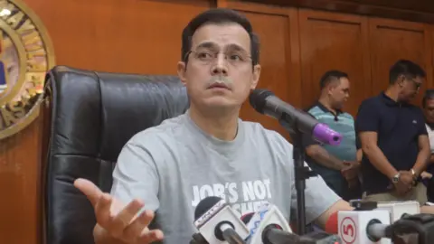 Isko Moreno Domagoso/Facebook Francisco Domagoso in a grey t-shirt, sitting at a wooden table. He is looking to the side and speaking to microphones resting on the table.