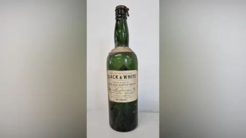Liversage Trust A composite image showing the empty Black & White Scotch whisky bottle, which is green glass with an off white label.