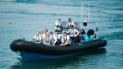 Robbie Dark The image shows a RIB carrying a number of people on board. Sophie is pictured at the front. They are all wearing lifejackets.