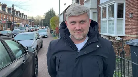 Phil Harrison/BBC A man with grey hair and wearing a navy coat is standing in a residential street beside a car.