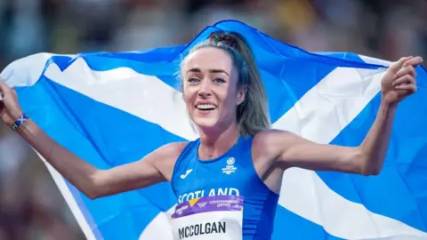 Getty Images Eilish McColgan wearing a blue running vest holding a Scotland flag above over her head and smiling at the camera after winning gold at the 2022 Commonwealth Games.