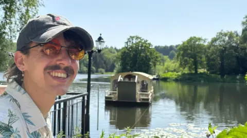 Jacob Edwards A man looking at the camera and smiling. He is wearing a cap and sunglasses. He is standing in front of a lake and there is a boat in the background.