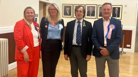 L-R: Lucy Stephenson, Arlene Maltman, Richard Honeycombe and Chris Rebindaine. All four are stood in a hall following an election count in Jersey. Stephenson, Maltman and Redindaine have rosettes on.