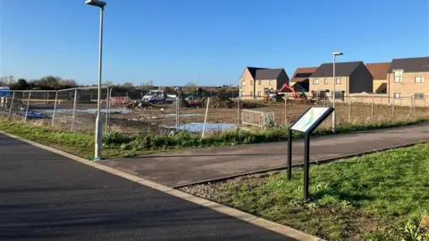 A plot of land partially surrounded by metal fencing in between a road and some houses. It is largely made up of mud. There are construction vehicles parked around its perimeter. 