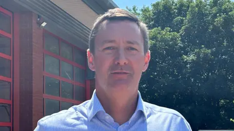 A man with a blue shirt standing outside a fire station. He has brown hair and light stubble