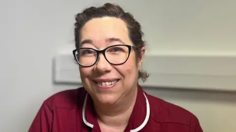Julie is sitting down in a room with a white background. She is smiling at the camera. Julie has brown hair and is wearing glasses with a black frame. She is wearing a dark red coloured nurse uniform.