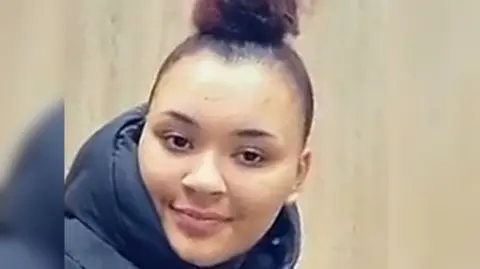 A girl reddish brown bun on top of head. She wears a black puffa coat and looks directly at the camera with a slight smile.