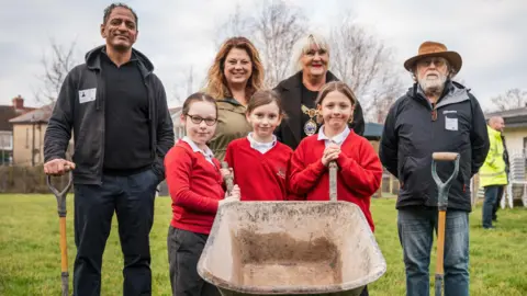 Sandylands Primary School Two men and two women are standing next to three schoolchildren who are holding a wheelbarrow. The school and the school field can be seen behind them.