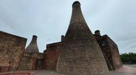 A series of brick-built pottery kilns are arranged under a grey sky.