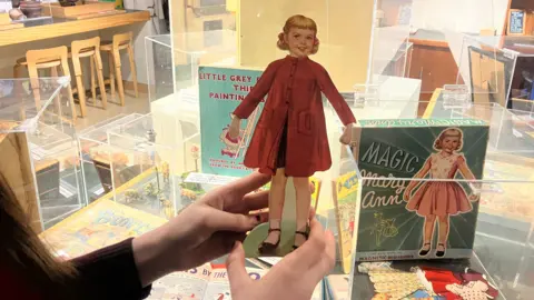 A cardboard cut-out doll is held in the hands of a girl. The doll has blonde pigtails and is wearing a red coat, black skirt and black shoes with white socks.