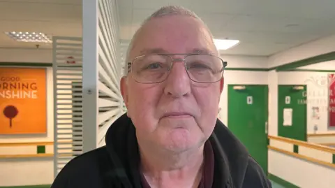 BBC/Louise Hobson John Daly is a man with short grey hair, glasses and a black fleece. He is stood in a large room.