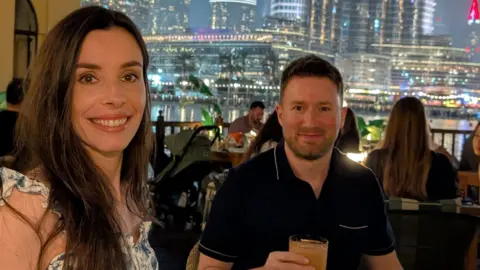 A woman with long dark hair is pictured with a man in a navy blue polo top with short dark hair and a beard. The man is holding a drink with a city landscape behind him at night.