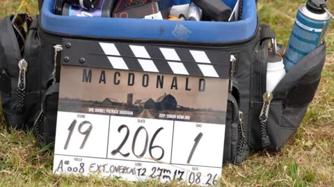 A black and white clapperboard sitting in long green grass on the set of MacDonald near Hexham. The board has the films logo on it and other details regarding the filming schedule.
