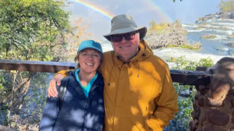 Roland and Shirley Cherry The couple are stood by a waterfall with rainbows appearing behind them as they stand against a fence. Mr Cherry has a hat on and a yellow jacket and is smiling with his arm around his wife who has a blue cap on and blue jacket and is also smiling.