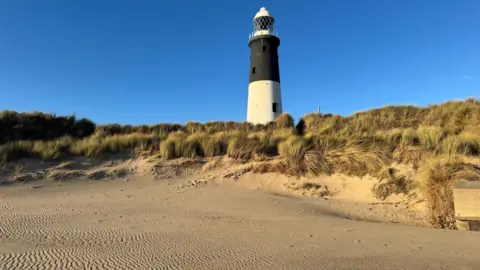 inspire23 A bright blue cloudless sky with a black and white lighthouse. There are sand dunes and marram grass in the foreground and a clean beach marked by ripples from the sea.