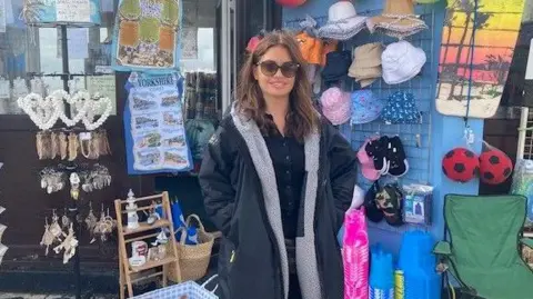 Sophie Walker, wearing a long black coat and sunglasses, stands outside her shop near Bridlington Harbour. She has dark brown hair. The shop has a variety of displays including seashells, hats and balls.