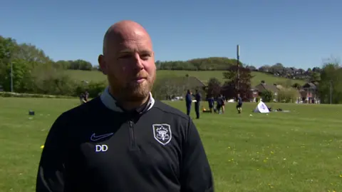 A man is wearing a black top including the initials DD. A field and other people are in the background, while trees and rooftops are in the distance. 