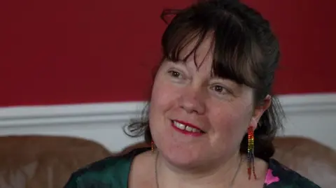 Santana Huckle sits on a sofa with a red wall behind her. She is wearing long slim rainbow-coloured earrings. 