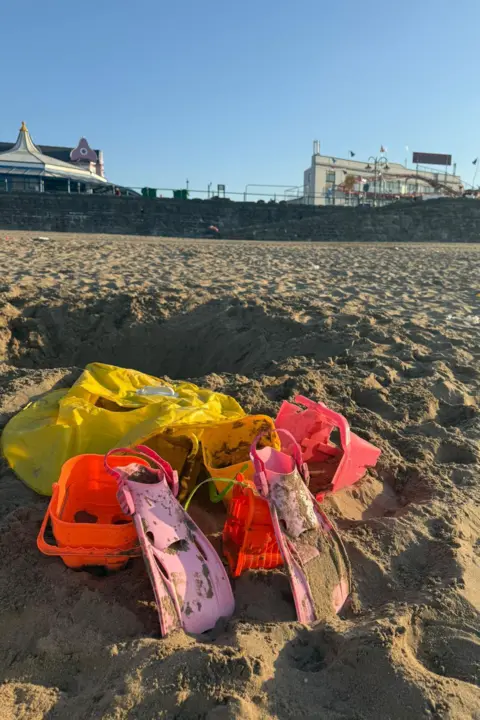 Lisa Wallace Pink flippers, orange and yellow sandboxes and a yellow lilo covered in sand. They are lying on the sand at the beach. 