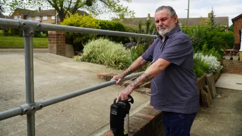 LOCAL DEMOCRACY REPORTING SERVICE Michael Partridge, wearing a blue shirt and black trousers, stands beside the kerb outside his house. He has an oxygen tank beside him, with a breathing tube running from the tank to his nose.
