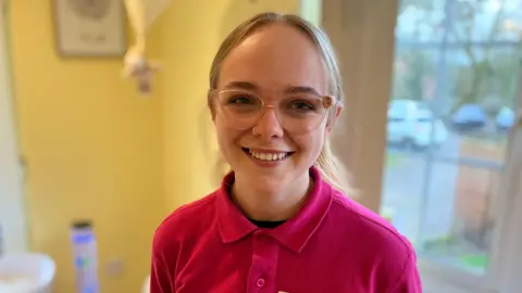 A woman looking at the camera and smiling. She is wearing a pink polo shirt.