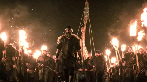 A bunch of men in Viking outfits and burning torches march in a line. Some are in modern dress too.