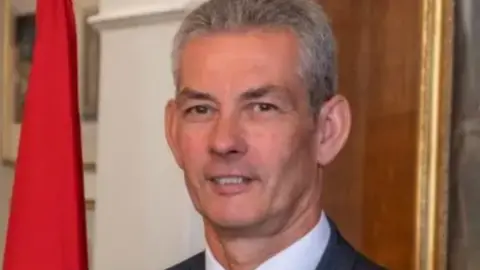IOM government Tim Crookall stands for a portrait. He has short, silver hair and thick eyebrows. He is standing in front of a Manx flag.