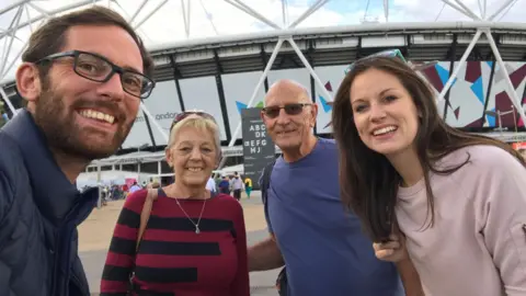 Peter Dunlop A man with a brown beard and glasses is taking a selfie with three other people in front of a stadium. The second person is a blonde woman with a burgundy jumper with black stripes, then a man with dark glasses and a blue t-shirt. On the right is a woman with long brown hair and a light-pink jumper. They are all smiling. 
