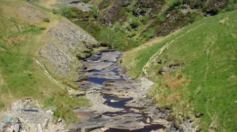 A dry river bed at Rhyd-y-Bedd in Conwy county.  A trickle of brown water is visible, making its way along the rocky river bed.  The grass on the river banks is starting to brown too.