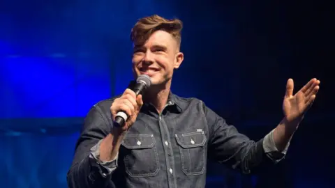 Getty Images Ed Gamble performing stand up holding a microphone wearing a denim shirt