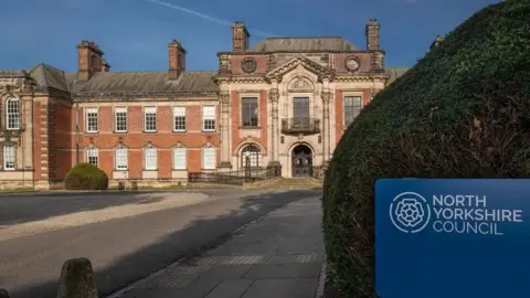The front of a manor house. On the right is a blue sign with white writing reading "North Yorkshire Council".