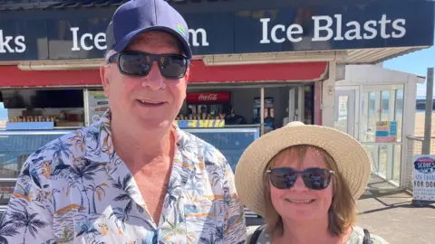 Julie Burton and Bernard Burton smiling at the camera. Bernard is wearing a white shirt with black, blue and orange palm trees. He is wearing a cap and sunglasses. Julie is wearing sunglasses and a cream wicker hat. They are both smiling at the camera. 
