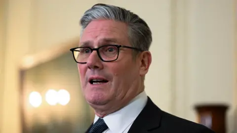 Reuters Britain's Prime Minister Keir Starmer looks off camera, wearing his customary black framed glasses and a dark patterned tie.