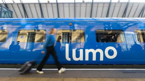 A blue train with the word Lumo on the side. A woman with a blue top and black trousers walks past the train pulling a suitcase