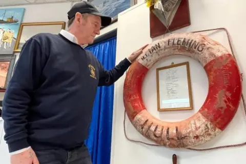 Kevin Shoesmith/BBC Jerry Thompson, un hombre de unos 60 años que viste una sudadera azul marino y una gorra de béisbol negra, está de pie junto a un aro salvavidas que muestra el nombre del barco pesquero Ian Fleming Hull.