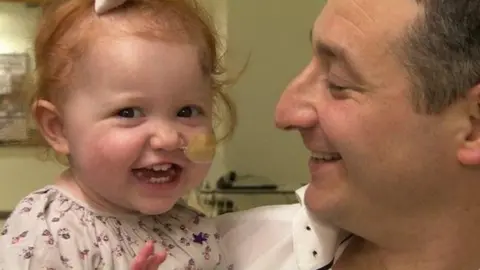 BBC A child with red hair smiles at the camera, she has a tube glued to her cheek, going into her right nostril. She wears a white top with purple flowers. She is held by a man in a white shirt who smiles at her.