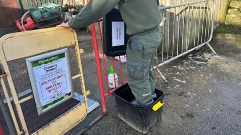 A man is standing in a bucket of disinfectant at a cleaning station. There is a sign that says: "Cleaning station, use footbath and spray all wheels."