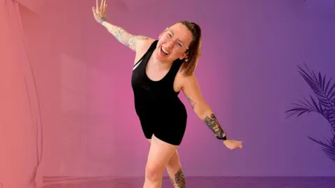 Movefit Mama A woman in a black vest and shorts standing with arms outstretched against a pink and purple background. She has tattoos on her arms and is wearing glasses. 