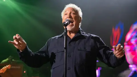 Tom Jones is standing in front of a black background singing into a microphone. He is wearing a dark blue shirt, and has grey hair and beard. 