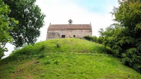 Getty Images A brown brick building with a small bell tower in the middle of it on top of a green hill, two sets of trees surrounding either side, blue skies with white cloud