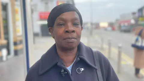 A woman wearing a dark coat and blue underneath is very prominent in the photo looking at the camera. She is near a road junction which is blurred in the background.