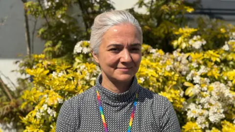 Zory Darman.  She is wearing a black and white turtleneck top and has long grey tied back hair. 