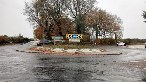 East Dunbartonshire Council A roundabout on a motorway on a rainy day with grey clouds. A black car is in the process of going around the roundabout. 