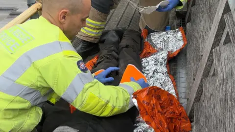 A firefighter is wrapping an orange piece of fabric around a dummy who is prone on the ground on top of the cathedral. The dummy is lying on a reflective foil blanket. 