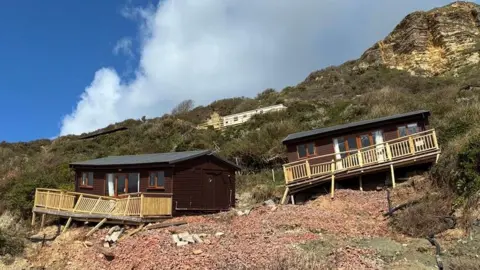 Two dark wooden beach chalets on a cliffside in Branscombe where they have become uneven and the decking has partially collapsed.