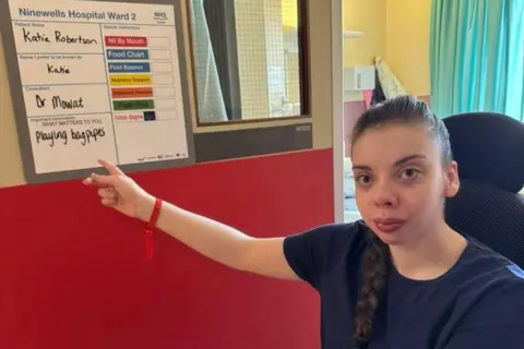 Katie Robertson A woman with a navy t-shirt and long brown hair in a plait pointing at a whiteboard in a hospital room.