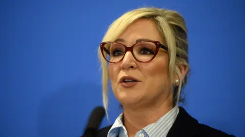 EPA Michelle O'Neill, wearing red-rimmed glasses, in front of a blue background