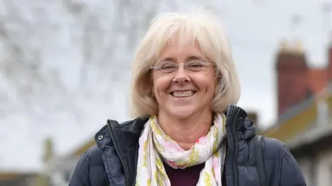 Getty Images A woman with bobbed light blonde hair and glasses, wearing a navy puffer jacket and white and pink floral scarf and smiling at the camera.